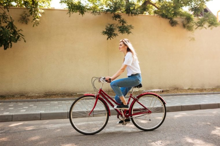 Beautiful girl with blond hair happily riding bicycle. Portrait of young lady in jeans and sunglasses on head having fun while riding on red bicycle along city streets