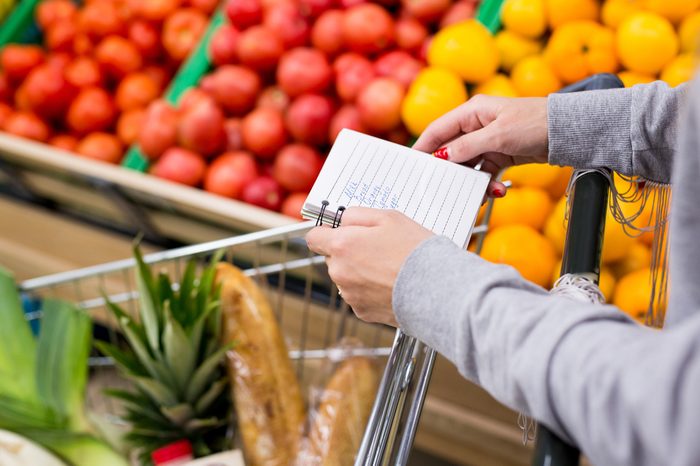 Woman with notebook in grocery store choosing vegetables, holding shopping list.