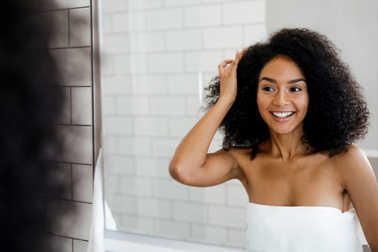 Young smiling woman examining her hair in the bathroom, looking into a mirror