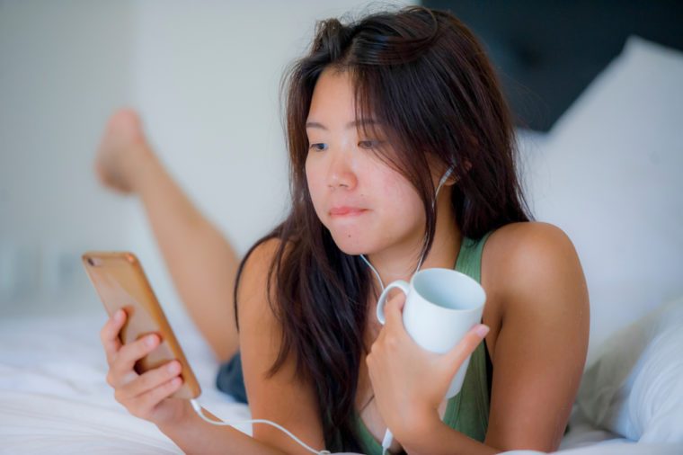 young beautiful and relaxed Asian Chinese woman with earpiece listening to music smiling happy lying on bed at home bedroom using internet on mobile phone drinking morning coffee cup