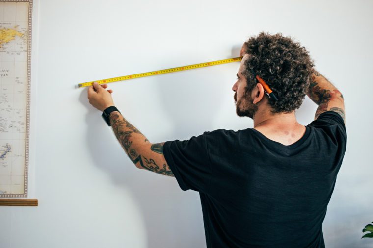 Hipster millennial man with arm tattoos and curly hair, small business owner, contractor measures wall to start reconstruction or adapt for painting, new home ownership 