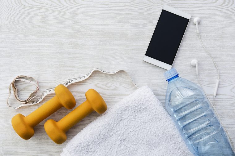 Orange weights, phone, headphones, measure tape, bottle of water and towel on wooden table. Fitness, sport time, flatlay