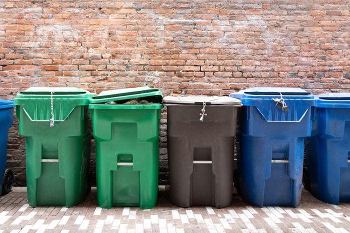 Row of green compost and clean green bins, beside blue recycling bins, with a brick wall background, and space for text on top