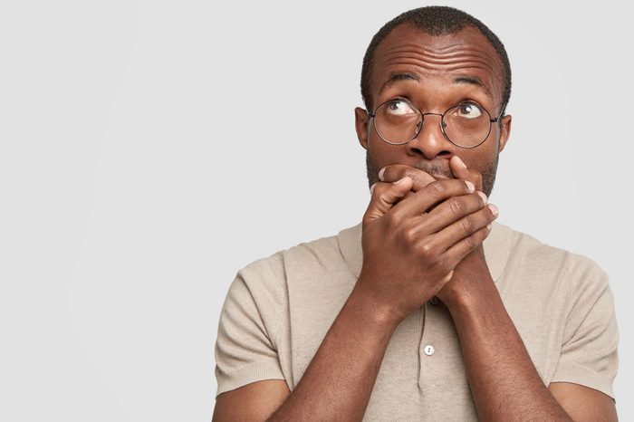Photo of dark skinned young male tries to keep silence, covers mouth and looks upwards with embarrassed expression, isolated over white background, blank copy space for your promotional text