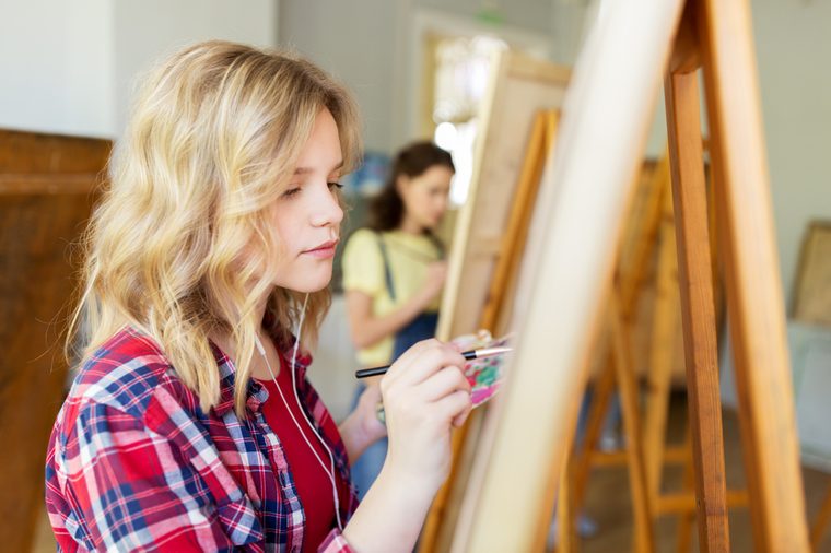 art school, creativity and people concept - student girl or artist with earphones, easel and paint brush painting at studio