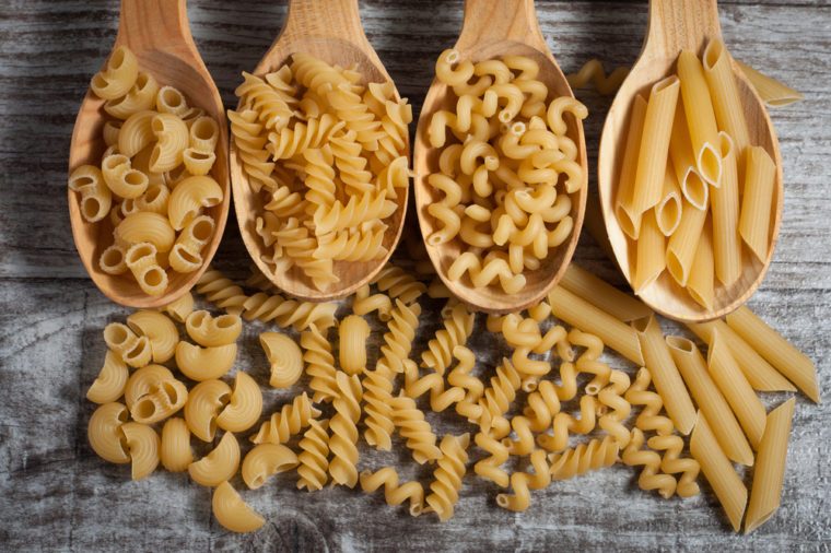 Various mix of pasta on wooden rustic background, sack and wooden spoons. Diet and food concept.