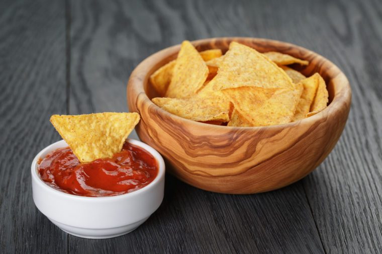 tortilla chips in olive wood bowl with tomato sauce on wooden table, selective focus
