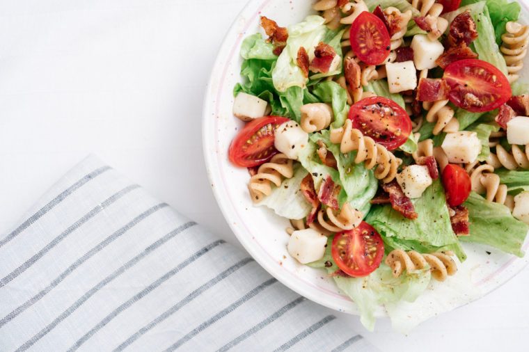 Overhead, close-up shot of BLT pasta salad with crispy bacon, grape tomatoes, green leafy lettuce and fresh mozzarella in white bowl with blue striped linen.