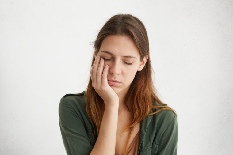 Cute woman having sleepy expression looking tired holding her hand on cheek closing her eyes with tiredness. Young woman having sad expression and toothache. People, problems, tiredness concept