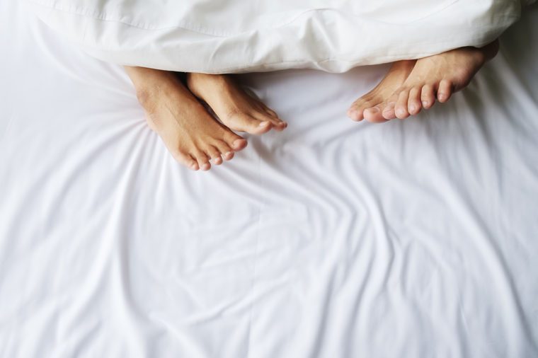 Feet of couple in comfortable bed. Close up of feet in a bed under white blanket. Bare feet of a man and a woman peeking out from under the cover.Top view with copy space (selective focus).