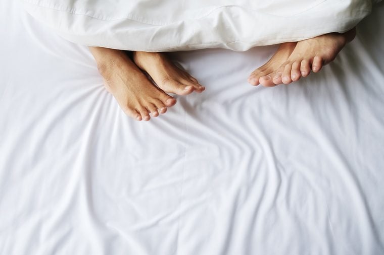 Feet of couple in comfortable bed. Close up of feet in a bed under white blanket. Bare feet of a man and a woman peeking out from under the cover.Top view with copy space (selective focus).
