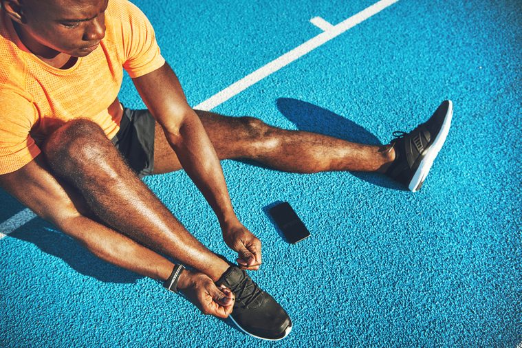 Focused young African male athlete in sportswear sitting alone on the lanes of a running track tying up his shoes before training