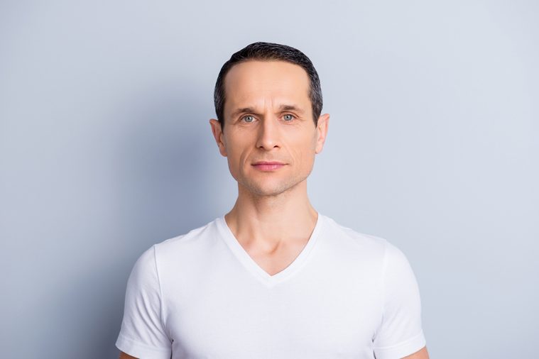Portrait of trendy, neat, shaven, brunet man in white t-shirt with serious expression looking at camera, isolated on grey background