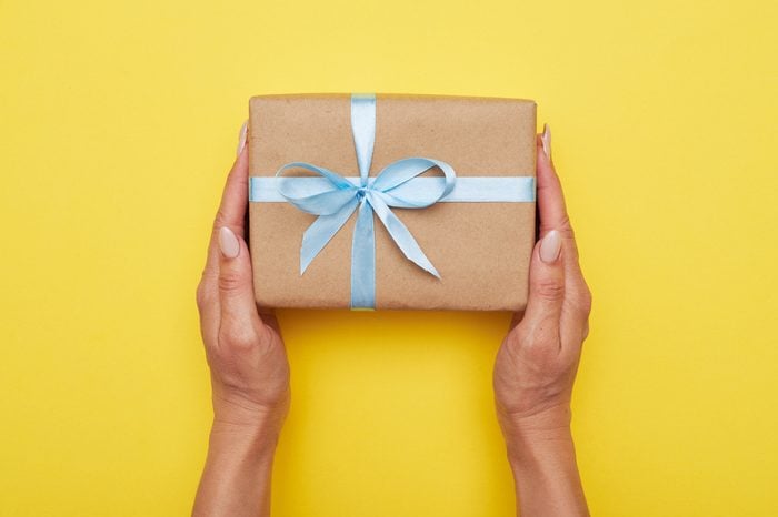 Flat lay of woman hands holding present with box decorated with blue ribbon on yellow background