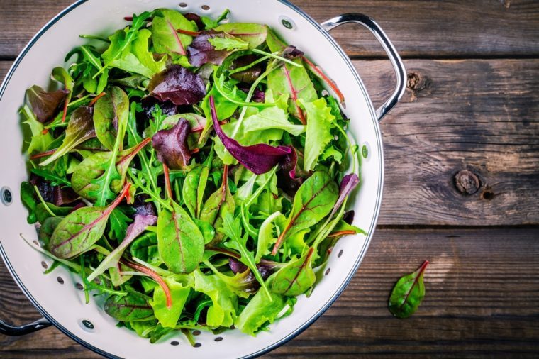 Fresh salad with mixed greens in colander on wooden background