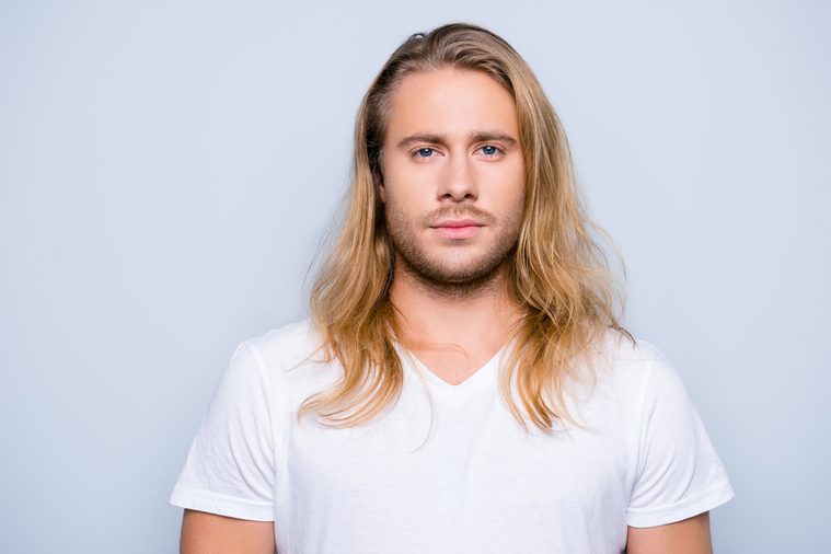 Portrait of handsome young serious confident young guy with blonde long hair in white tshirt, isolated on grey background