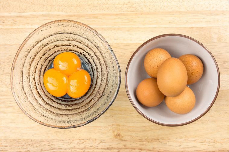 Raw eggs in ceramic bowl and egg yolks in glasses bowl on wooden table, top view