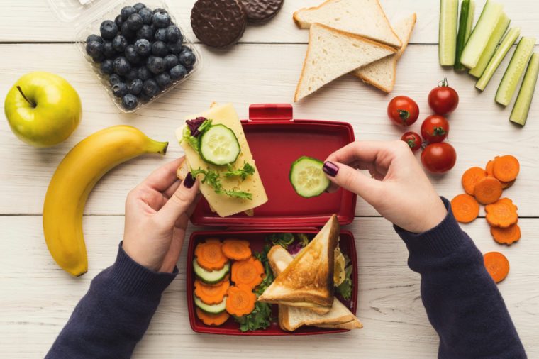 Preparing healthy vegetarian snacks on white rustic wood. Female hands making sandwiches and putting into take away plastic lunch box, top view. Eating right, picnic and food storage concept
