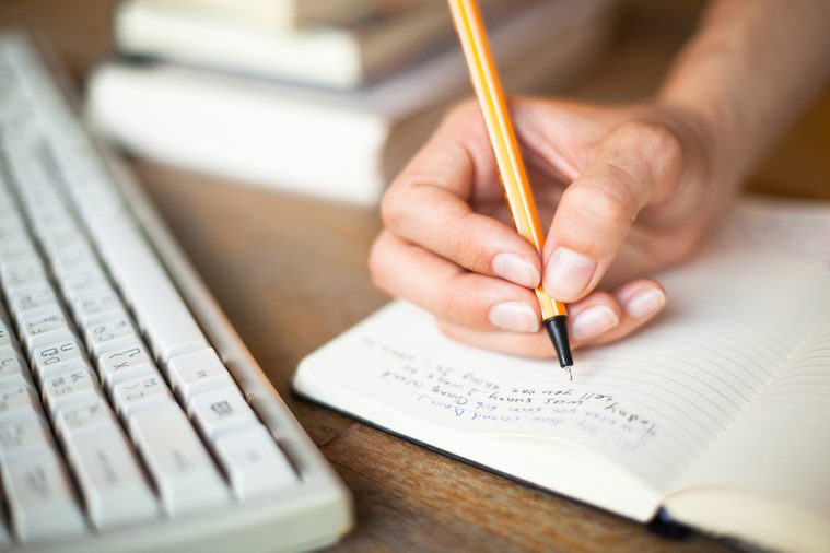 Photo of hands writes a pen in a notebook, computer keyboard and a stack of books in background