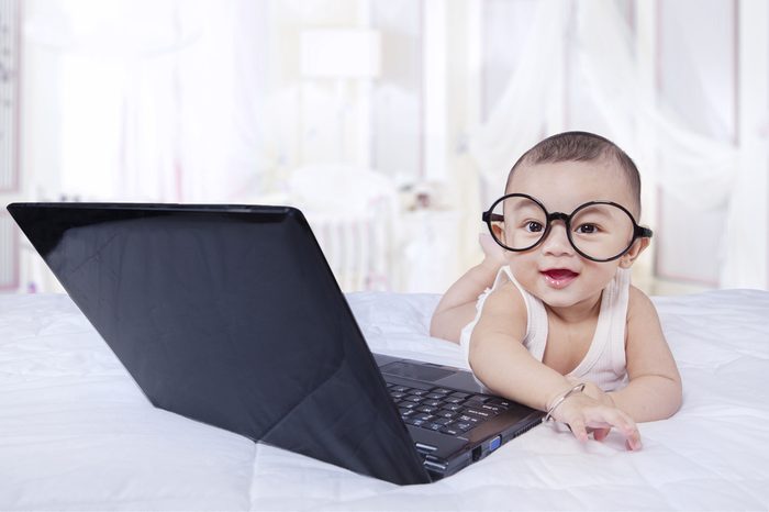 Portrait of beautiful little baby boy lying on the bedroom with glasses and laptop