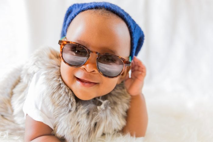 Super cool baby. Hipster baby in fur vest and sunglasses lies on a white bed in a room with curtains. Baby smiles while putting on sunglasses.