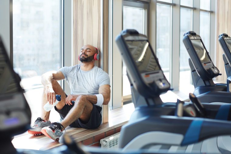 Portrait of modern trendy sportsman sitting in sunlight by window listening to music in big headphones with eyes closed, resting after gym workout behind row of treadmills