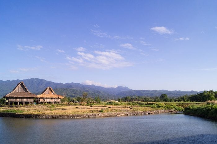 A scenic of view of the local landscape in Bagac, Bataan, Philippines. Two wooden houses stand along a river. Mountains surround the area.