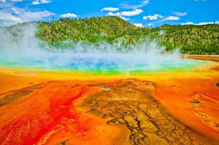 Beautiful cerulean geyser surrounded by colorful layers of bacteria, against cloudy blue sky.