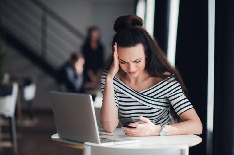 Portrait of stressed office young woman holding a cellphone in hands in a cafe, looking at the screen with cross face expression, mad at stressful texts and calls.