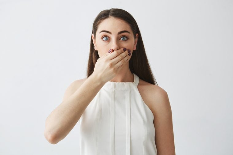 Portrait of surprised young brunette girl covering mouth with hand looking at camera over white background.