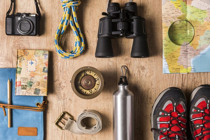 Overhead view of travel equipment for a backpacking trip on wooden floor. / Items include rope, belt, canteen, compass, map, camera, binoculars, journal, boots. Time to travel concept.