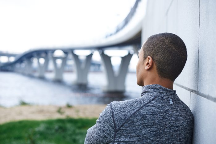 Rear view of thoughtful African American male runner in stylish sports jacket leaning on wall outdoors, looking into distance at sea, relaxing mind or meditating after running exercise in the morning