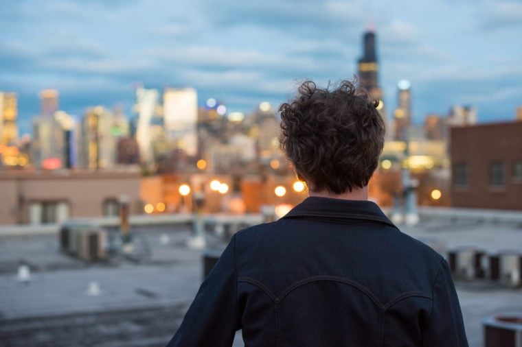 Attractive, forty something man, with long, curly brunette hair, on a rooftop, in the city, with cityscape in the distance. Dusk and sunset lights. Staring into the distance. Contemplating the future.