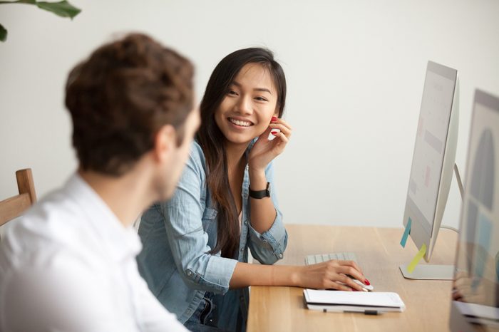 Smiling attractive asian woman talking to male colleague at work sharing office desk with desktops, friendly multiracial coworkers interns having pleasant fun conversation at workplace together