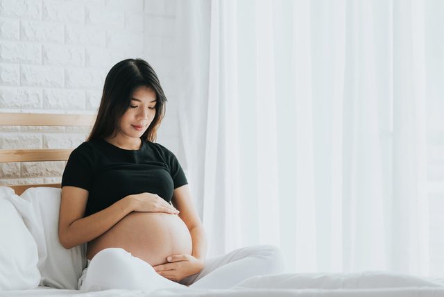 Happy Asian pregnant woman touching her belly with care in bedroom. She can??t wait to be mom.