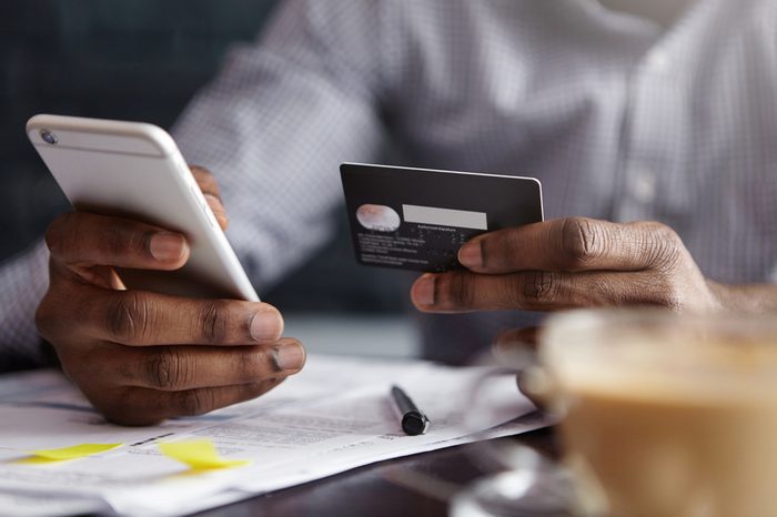 Cropped shot of African-American businessman paying with credit card online making orders via Internet. Successful black male holding plastic card making transaction using mobile banking application