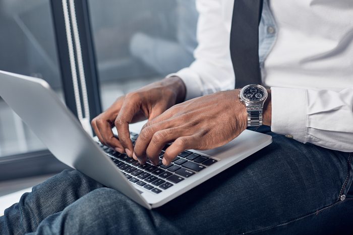 Important task. Close-up of hands of young african manager is sitting and holding laptop on his knees while typing on keyboard
