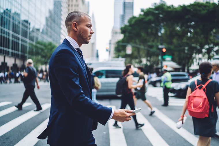 Side view of confident businessman passing crosswalk in modern district of megalopolis choosing direction, serious male entrepreneur crossing street with crowd getting to office building by foot