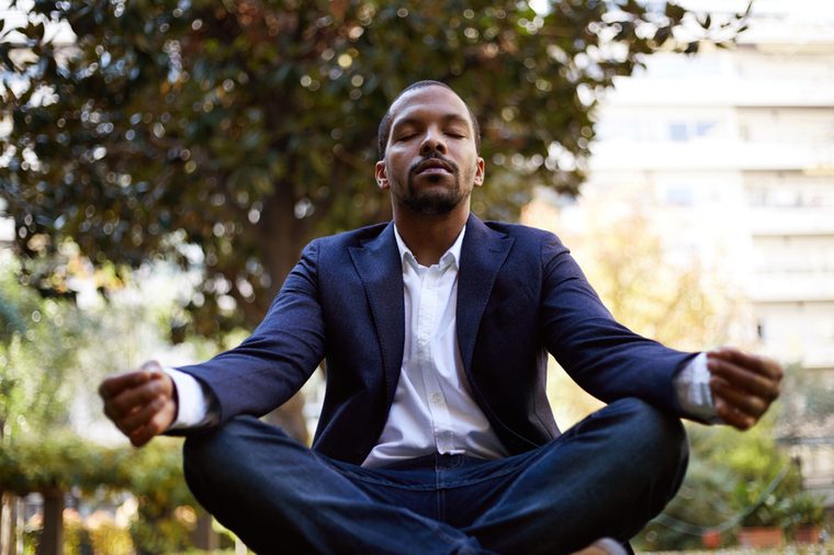 Young american african businessman in informal clothes meditating in lotus pose taking a deep breath outside corporate office.Business yoga and stress free environment.Peace of mind concept.Blurred