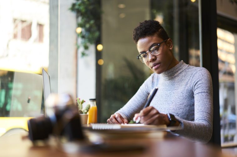 Concentrated afro american experienced woman journalist in glasses creating article working in coffee shop, skilled young female graphic designer making sketch in notebook for new startup project 