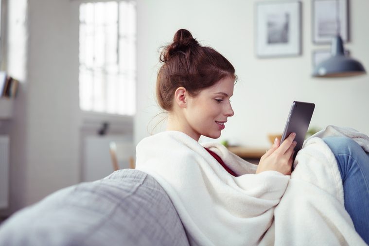 Happy young woman enjoying an e-book on her tablet computer as she relaxes with her feet up on the couch wrapped in a warm blanket, side view with copyspace