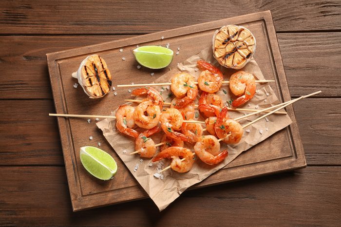 Board with delicious fried shrimp skewers, lime and garlic on wooden background, top view