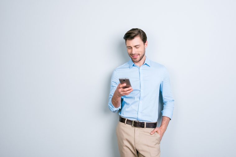 Portrait with copy space of attractive, cute man holding smart phone in one hand and another one in pocket, checking email through wifi, 3G internet over grey background