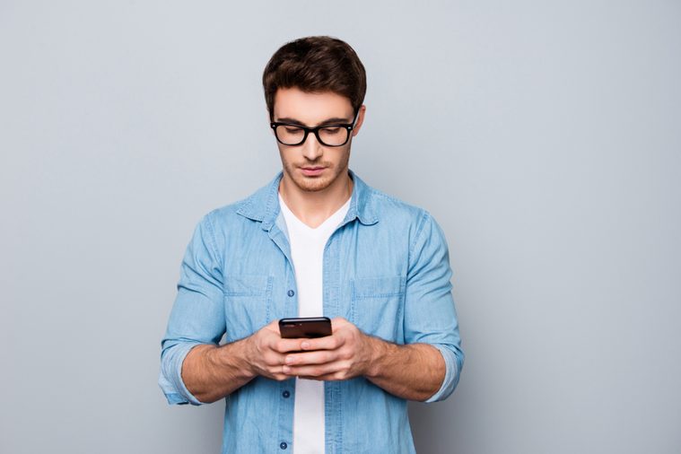 Portrait of concentrated, busy, trendy, modern, brunet guy with stubble, hairstyle having smart phone in hands, checking email, using 3G, wi-fi internet, searching contact, isolated on grey background