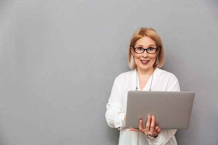 Smiling middle-aged blonde woman in shirt and eyeglasses holding laptop computer while looking at the camera over grey background