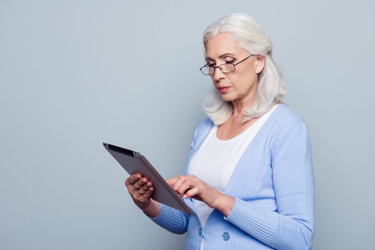 Portrait of pretty, charming, aged woman with holding tablet, using wifi internet for work, checking email, standing over grey background