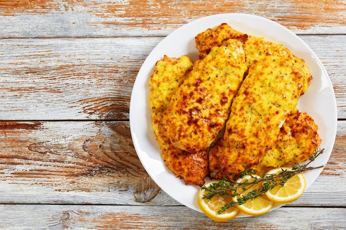 delicious Baked Chicken breast coated with melted emmental cheese and Whole-grain mustard on white plate with thyme and lemon slices, on old wooden table, view from above