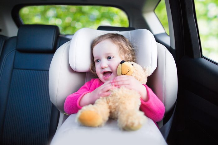 Cute curly laughing and talking toddler girl playing with a toy bear enjoying a family vacation car ride in a modern safe vehicle sitting in baby seat with belt having fun watching out of the window 