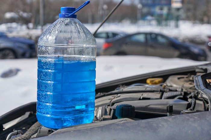 Bottle with non-freezing liquid for windshield washer standing on the engine of a car in front of a road with cars in winter outdoors