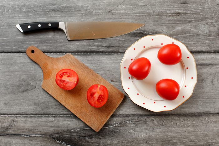 Table top view on three tomatoes on white porcelain plate with red dots, cutting board with tomato cut to half next to it, chef knife also laying on the gray wood desk.